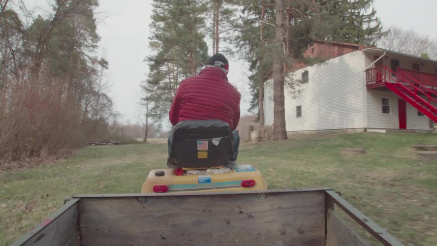 A man rides a lawn tractor trailer in a yard near a house