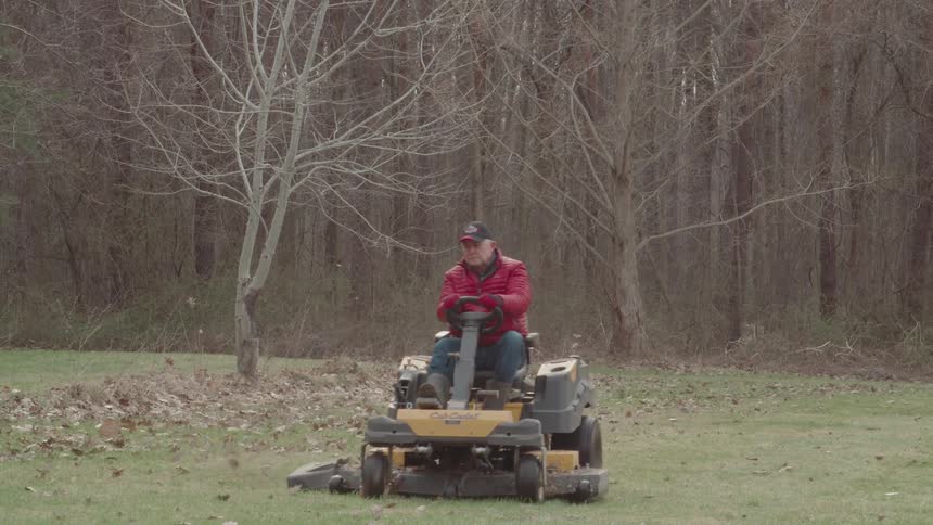 A man wearing a red jacket and a baseball cap drives a yellow riding mower across a grassy lawn covered in fall leaves with a forest of bare trees in the background