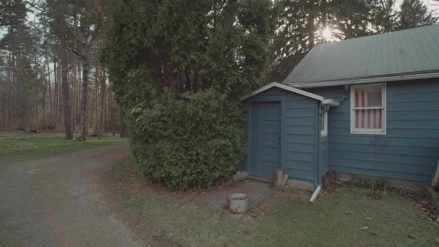 A drone shot captures the exterior of a rural home the scene is a mix of a wooded forest and a dirt driveway that leads up to the blue house an american flag hangs in one of the windows