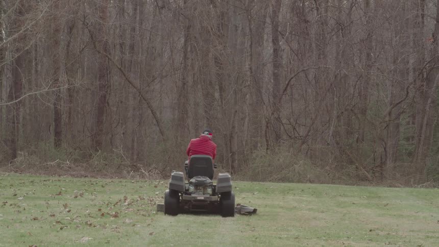 Based on the video a man in a red jacket and baseball cap is on a lawnmower he is seen driving the lawnmower away from the camera mowing the grass