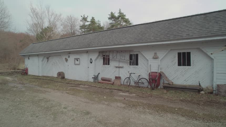 A view of a long white barn with a dark roof and a bicycle wheelbarrow and various other tools and objects lying around outside