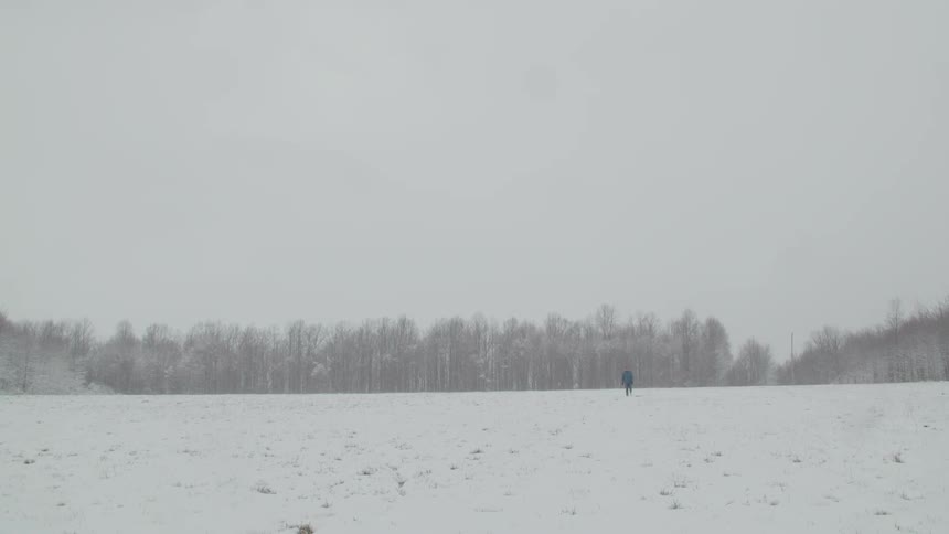 Adult man wearing a baseball cap and a blue jacket walking on a flat area covered by snow with a forest on the background outdoors during day time