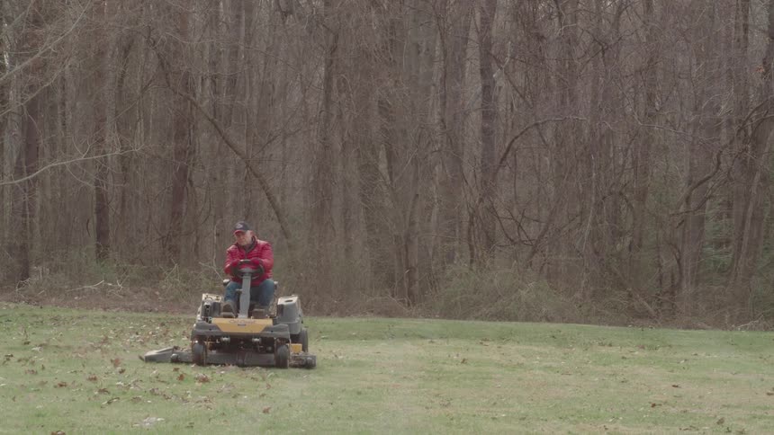 A man in a red jacket rides a lawn mower across a grassy field clearing leaves near a wooded area
