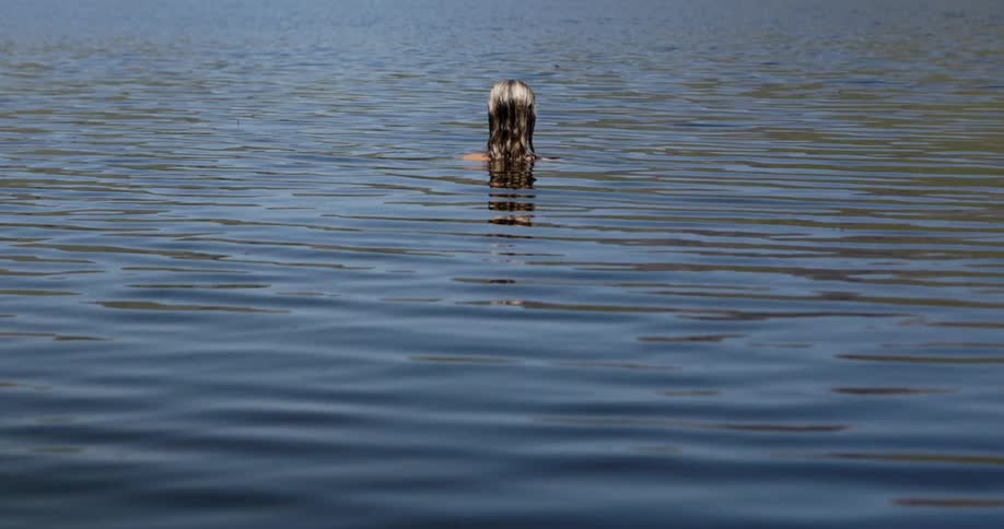 A blonde person is submerged in water with only the top of their back and their wet hair visible above the surface the video is a static medium shot showing the persons back and the ripples of water around them while their hair hangs down in the wate