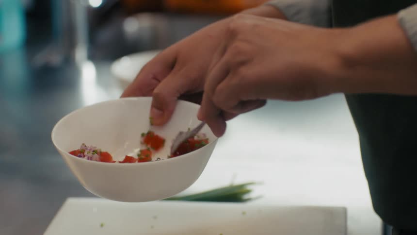 The video shows a chef using a spoon to mix a small bowl of diced ingredients including what looks like tomatoes and red onion the bowl is white and the chefs hands are visible against a blurry kitchen background