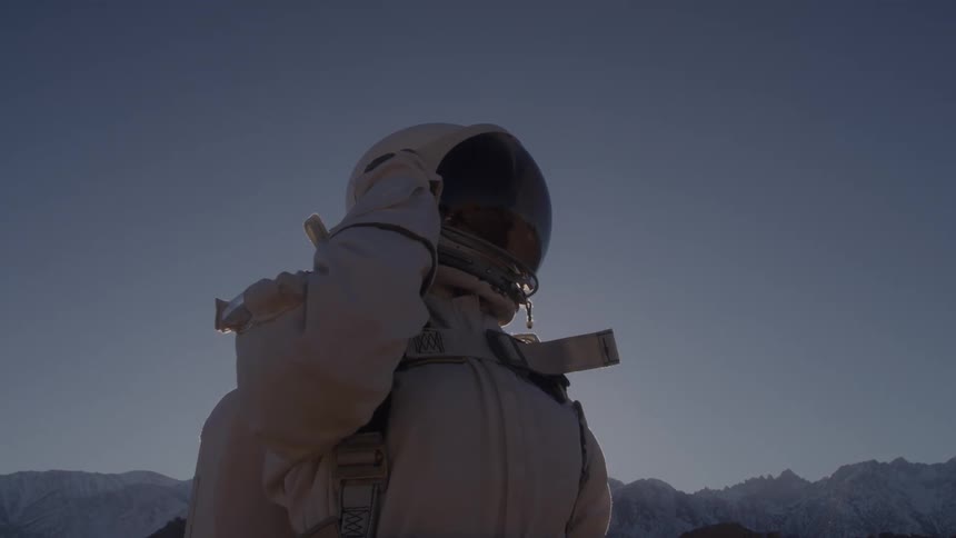 A person in a space suit stands outside and raises a hand to the side of the head