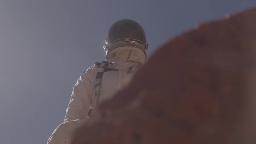 A person in a space suit stands on rocks under a blue sky