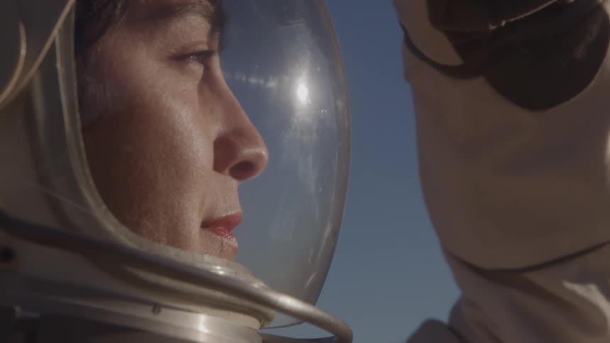 A close up portrait of a woman in a space suit looking to the front and a blue sky in the background
