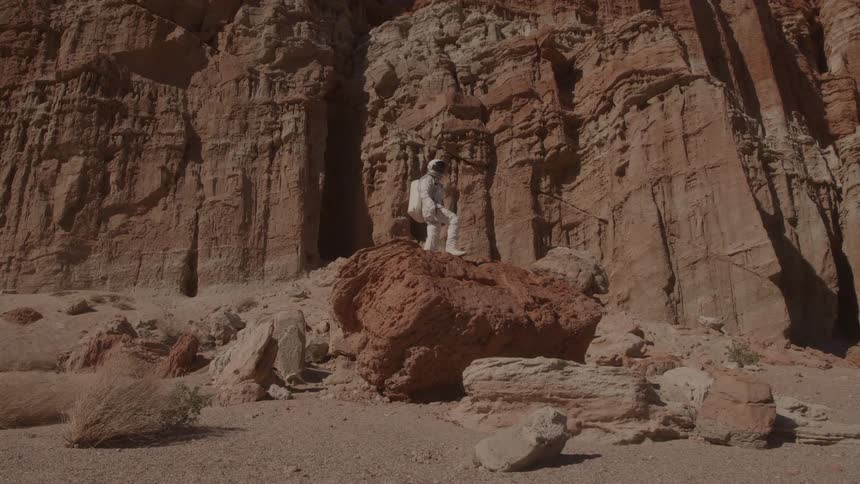 In a wide outdoor shot a person in a white spacesuit stands on a large reddishbrown rock surrounded by the towering striated walls of a canyon the scene has a reddishbrown dry and rocky appearance