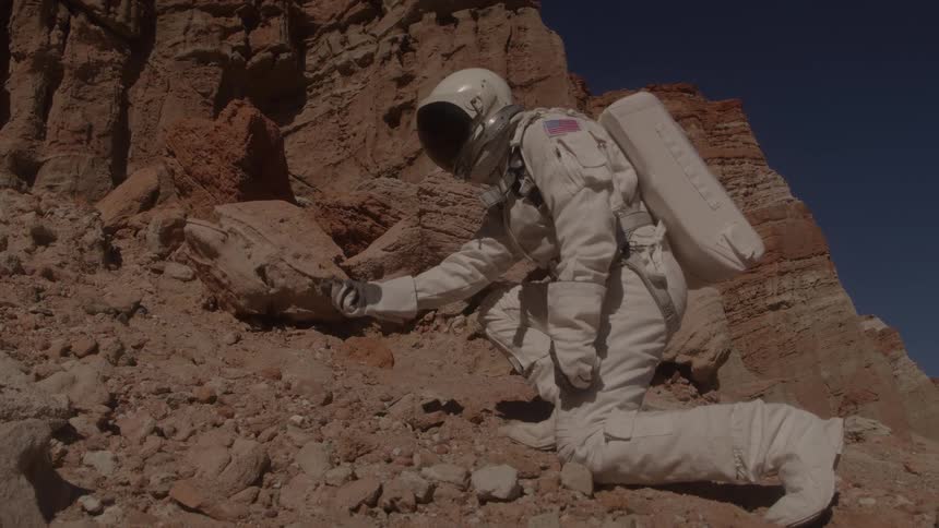 An astronaut in a white spacesuit is kneeling on a rocky brown landscape with a cliffside in the background looking at a small handheld device