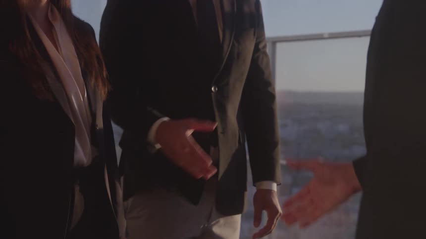 Three people including a woman and two men all wearing suits exchanging handshakes in a highrise office building at midday
