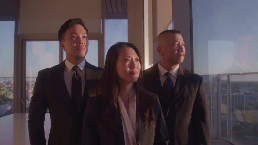 A midshot shows three business professionals in a line all looking toward the right of the frame the woman in the center faces the camera while the two men on either side look to the side