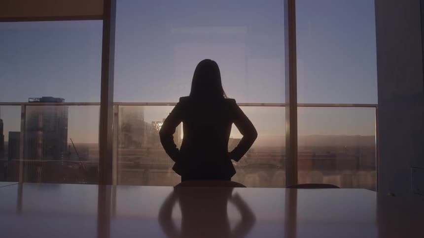 The woman is standing in the conference room watching the sunrise out the window