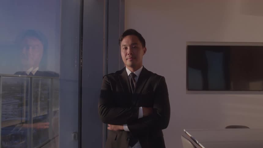 A midshot captures a man in a business suit standing indoors looking at the camera with a neutral expression he is positioned to the right of a large window which reflects his image