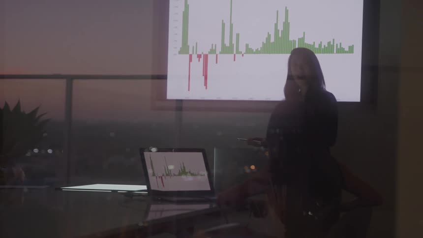 A wide shot from a distance shows a person silhouetted against the light standing in a dark office they are pointing at a graph on a large projection screen while facing a table with two laptops which are also displaying graphs