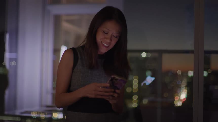 A woman stands indoors at night holding her phone and smiling as she looks down at the screen the background is a large window revealing the blurred illuminated cityscape