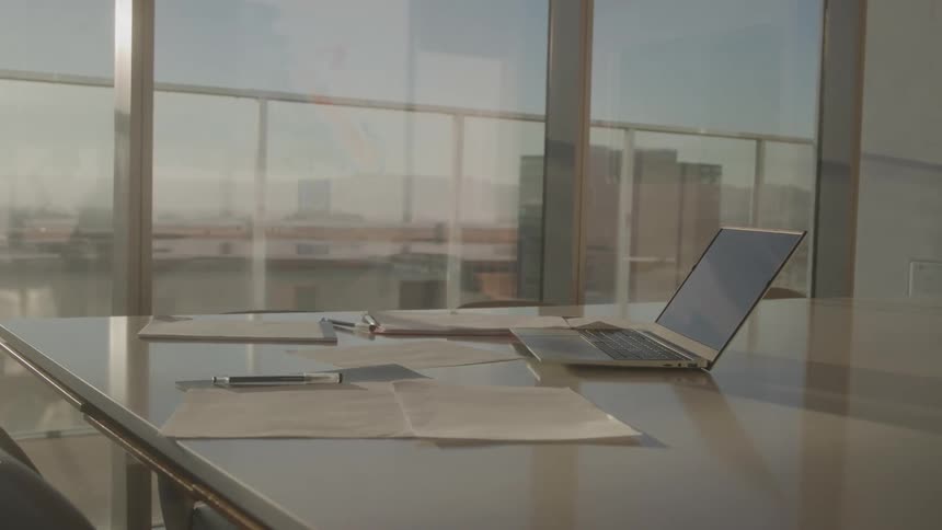 A closeup shot focuses on a clean empty conference table with a laptop and some papers arranged on it the background shows a bright hazy urban skyline through large office windows