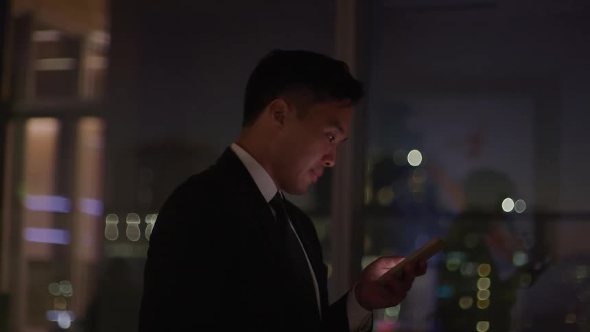 An asian man in a suit is looking at his cellphone at night in an office setting