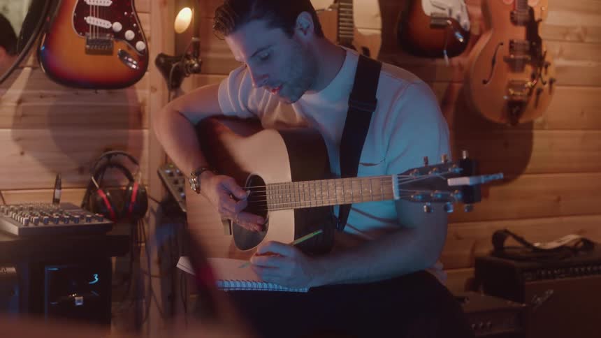 A man wearing a white shirt is sitting and playing an acoustic guitar while writing in a notebook in a music studio with several electric guitars hanging on the wooden wall
