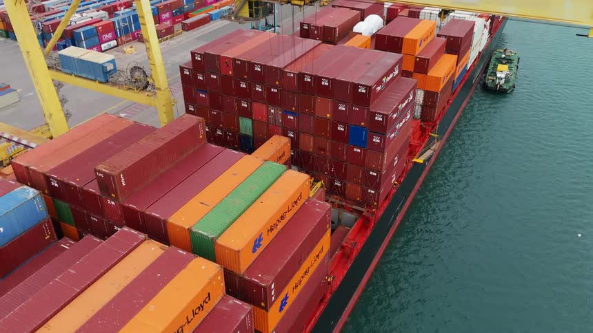 Piles of shipping containers by the sea on a cargo depot with a crane above them and a small boat beside them
