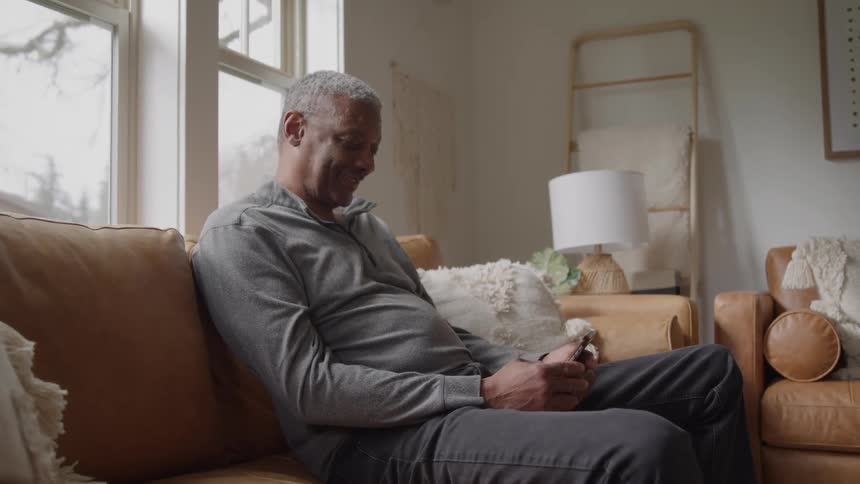 A senior man wearing a gray shirt and black pants is sitting on a brown couch in a sunlit living room and looking down at a phone he is holding