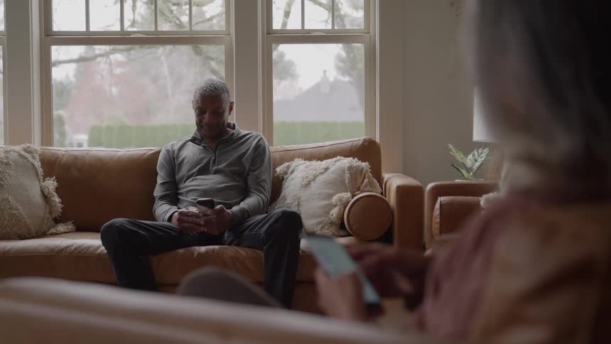 Based on the video a man is sitting on a couch and looking at his phone another person is sitting on a different couch also looking at their phone