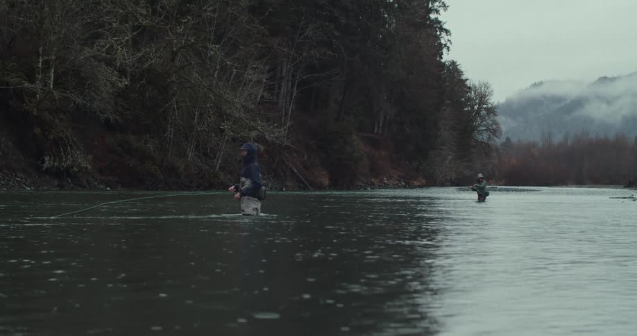 Two people are fishing in a river the water is murky and the trees on the riverbank are bare the sky is grey and there are mountains in the distance