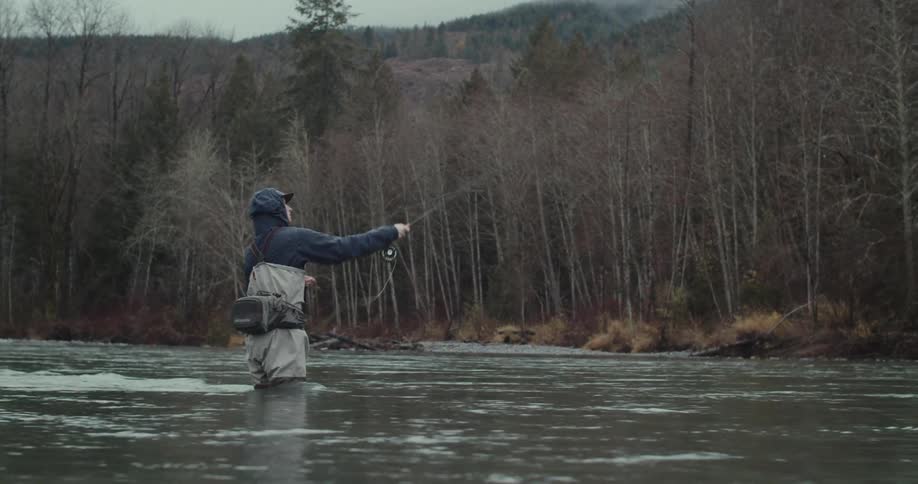 A person stands in a river and casts a fishing line with a rod surrounded by trees and mountains