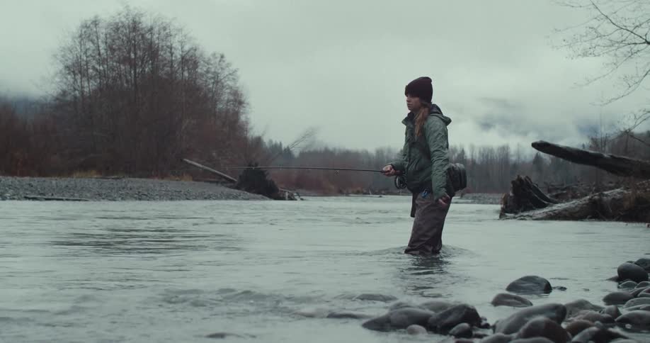 A young woman standing up to her knees in a river holding a fishing rod pulling on the fishing line then throwing it back