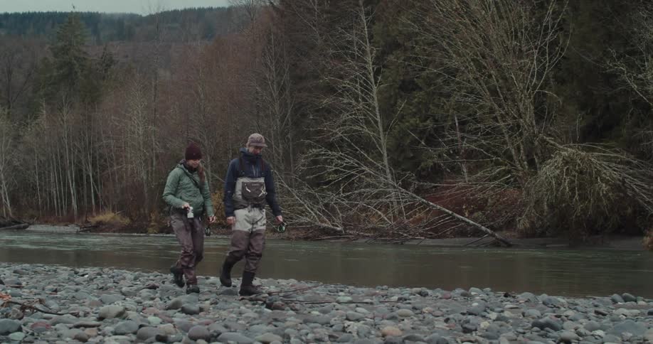 Two people walk on a rocky riverbank holding fishing rods and wearing waders with a river and trees in the background