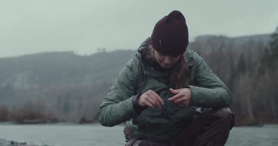 A woman wearing a puffy green jacket and a beanie is crouching near a river with a focused expression as she ties a fishing line on a cloudy day