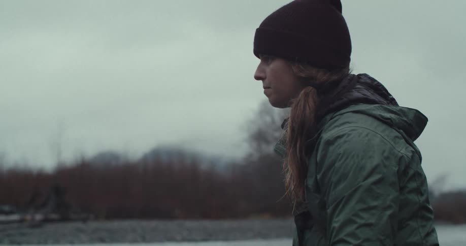 A closeup side profile shot shows a person wearing a beanie and a green jacket they are holding a fishing rod and looking intently at the water the background is a blurry river and a treeline under an overcast sky