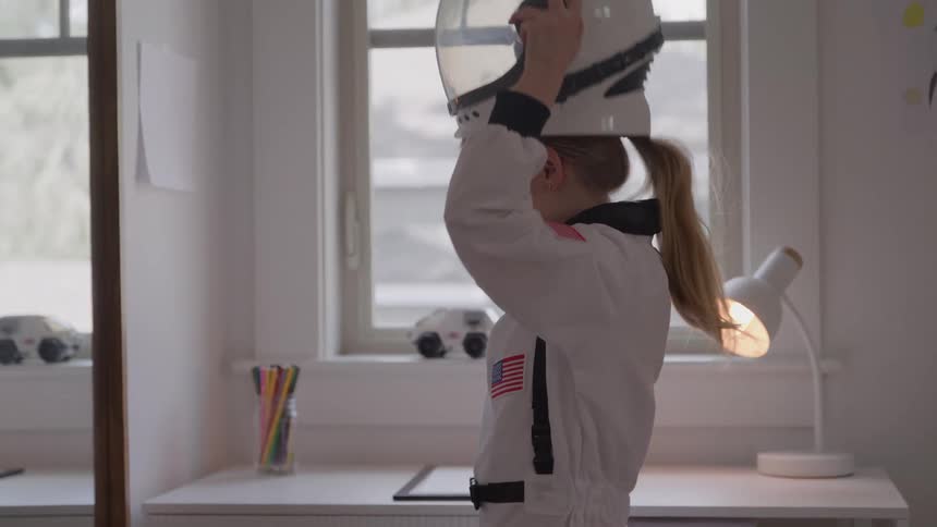 A young girl in a white astronaut suit with a united states flag on her arm is standing in her bedroom and puts on a large white astronaut helmet