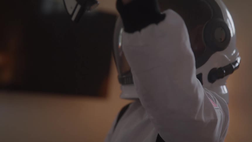 A close view shows a young girl in a white astronaut suit and helmet playing with a toy shuttle in a dimly lit room with an american flag patch on her sleeve