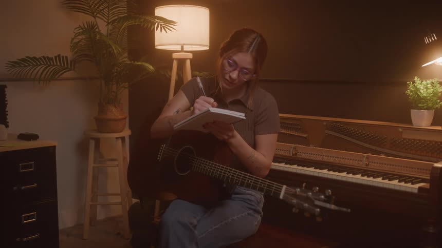 In a warmly lit music room a young woman with glasses holds a guitar while sitting at a piano and writing lyrics in a notebook