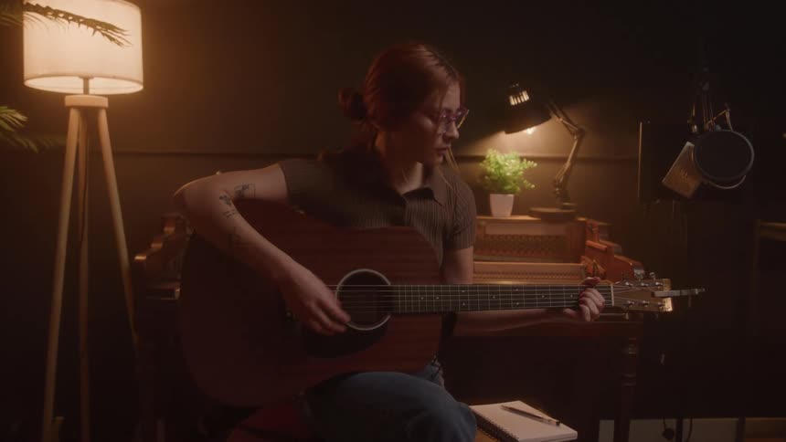 A young woman sitting with her back to a piano while playing acoustic guitar with a pen and notebook on a bench next to her