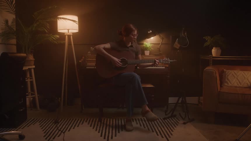 A young woman is sitting in a chair playing an acoustic guitar she is in a dimly lit room with a floor lamp next to her and various pieces of furniture and plants are visible