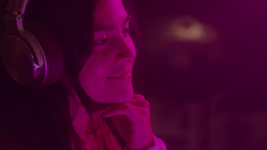 A close side view of a young woman with a long dark hair wearing large headphones and smiling as a pink light shines on her in a dark room