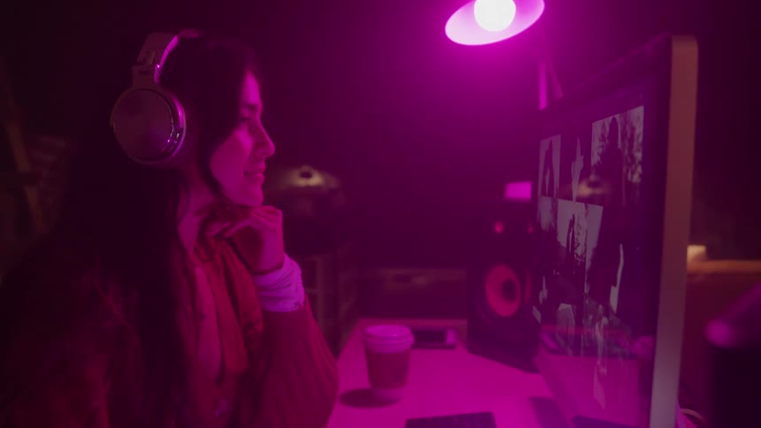 A young woman wearing headphones is shown from a side angle sitting in a dimly lit room with pink and purple lighting she is looking at a computer monitor that shows a video conference with other people she is holding a pen in her hand and seems to be