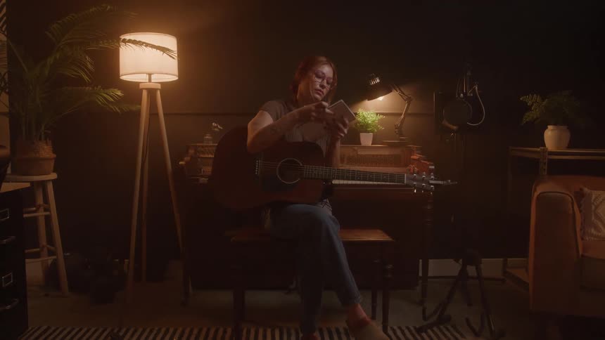 A woman wearing a brown shirt and jeans sits in a dimly lit music studio playing an acoustic guitar and writing notes in a notebook with a glowing floor lamp and a vintage piano in the background