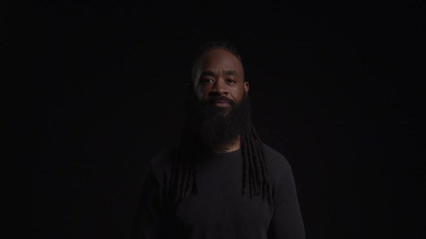Black man with a bald head and long black beard dressed in black clothes standing in a dark room