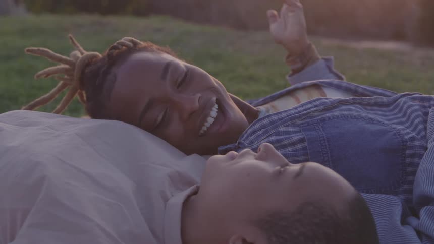 An affectionate couple lies together on a blanket in the grass at sunset smiling and looking lovingly at each other