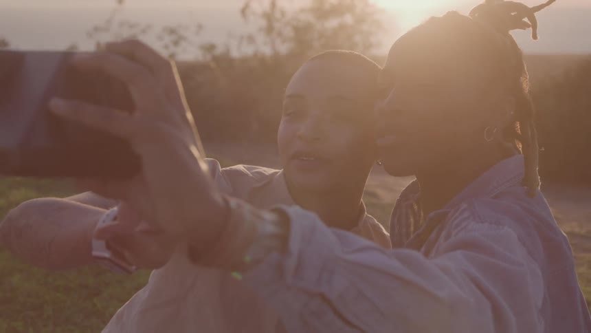 Two young people one with their back to the camera are taking a selfie with a tablet they are standing outdoors and the setting sun behind them creates a warm hazy glow