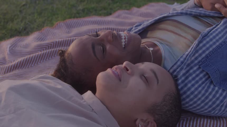 A close view of a woman with short blond hair and a woman with dark curly hair laughing happily as they lie together on a striped blanket in a park on a sunny day