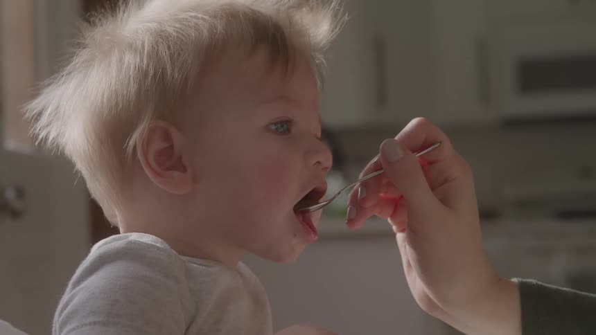 A close view shows an adults hand feeding a young baby with a spoon in a bright kitchen with white cabinets in the background