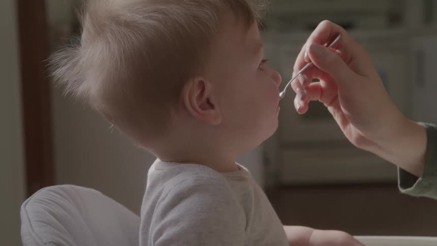 A close side view of a parent feeding a young baby with a spoon in a high chair in a kitchen with soft natural lighting