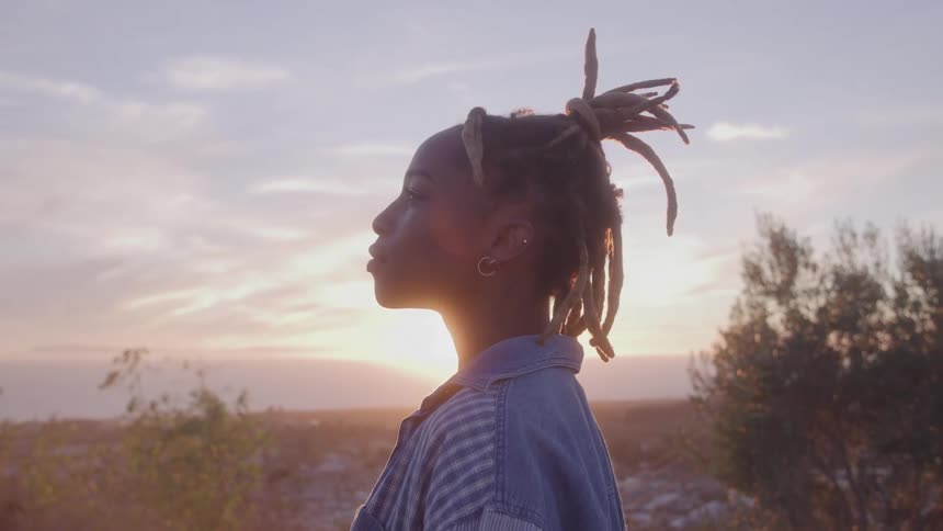 A profile view shows a young woman with dreadlocks and a jean jacket standing outside and looking out at the sun setting over the ocean