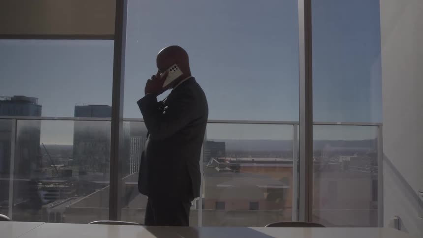 A silhouetted businessman in a suit paces back and forth while talking on the phone in front of a large window with a city view