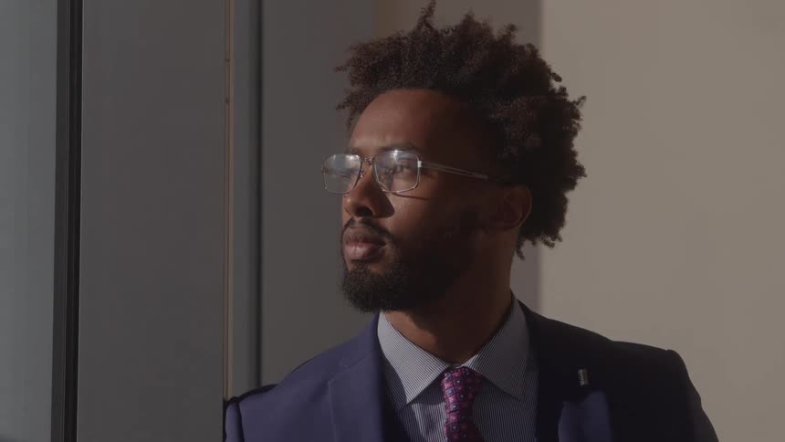 Young black man with afro hair wearing glasses standing beside a wall indoors at midday