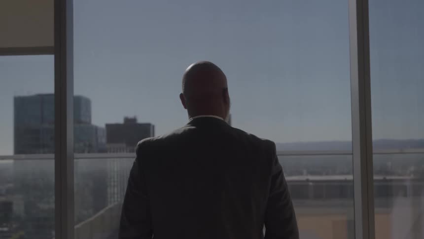 A man in a suit stands with his back to the camera and looks out a window at a city